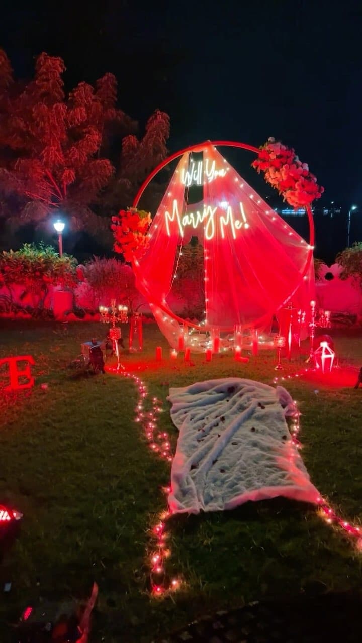 A proposal set up on the lawn after dark, a lit hoop hung with flowers and a neon sign reading will you marry me, with candles along a path in the grass.