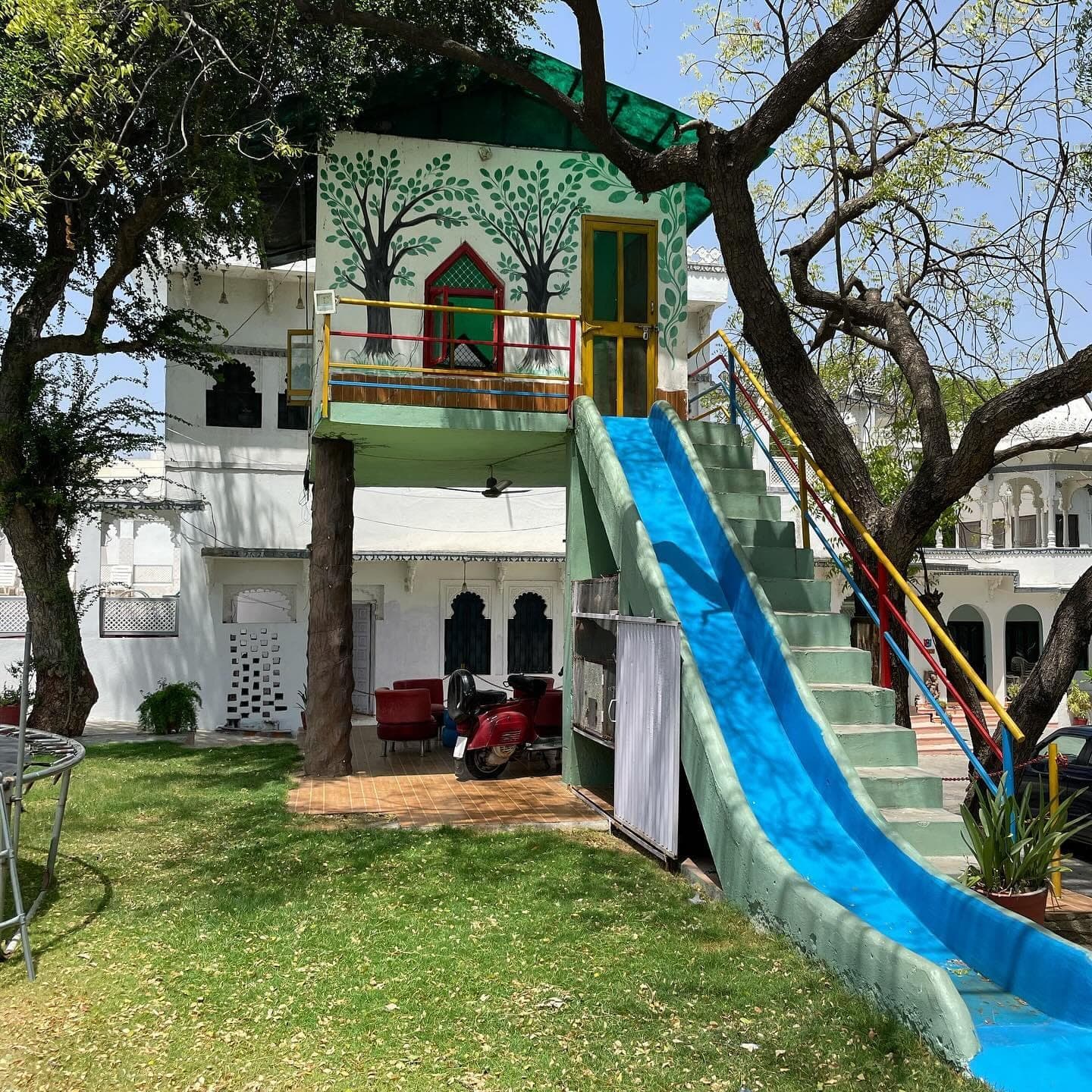 A painted tree house standing among the trees on the lawn, a blue slide running down from its balcony and steps climbing to the door.